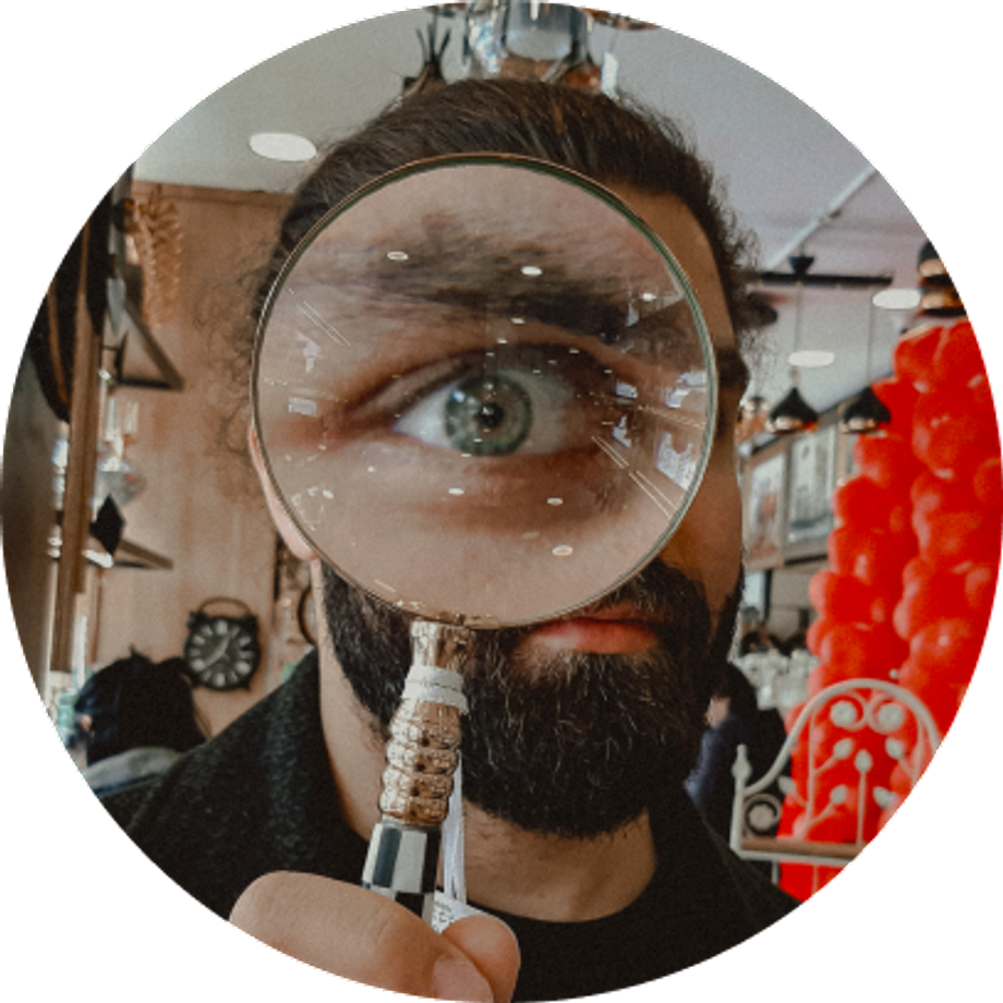 A White bearded man looking at the camera through a magnifying glass